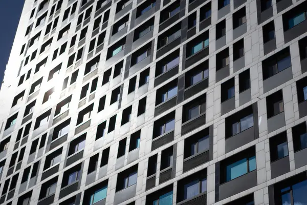 Açık gökyüzünü yansıtan güneş ışığında pencereleri olan yüksek bir apartmanın ön cephesi. Çok katlı yapı, öğlenden sonra aydınlıkta doku, renk ve mimari ritmin bir karışımını gösteriyor..