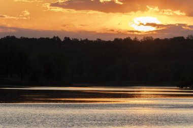 Güzel bir günbatımı, durgun nehrin üzerinde solan ışığı yansıtan altın ve turuncu renkler bırakır. Siluetli ağaçlar kıyı boyunca uzanır. Huzurlu ve nefes kesici bir manzara yaratır..