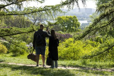 2025.05.01, Kyiv, Ukrayna: Güzel bir parkın kenarında duran iki kişi, çarpıcı şehir manzarasını telefonla çekiyor. Etrafı yeşillik ve açık gökyüzü ile çevrili, sakin manzaraya ve kentsel ufka hayran olmak için durdular..