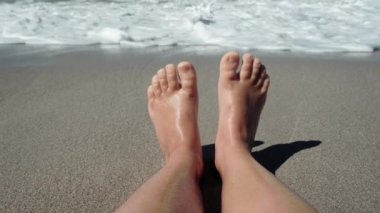 The feet of a child resting on a sandy beach against the backdrop of the sea. Waves with foam rolling up on the childs legs. Family vacation with kids. High quality 4k footage