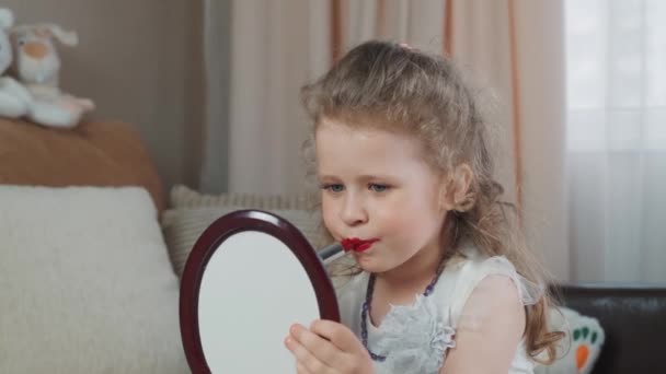 快乐的小女孩从妈妈那里借来的鲜红色口红涂在嘴唇上 并在镜头前亲吻 有趣的婴儿自己化妆 愉快地看着镜子里的自己 高质量的4K镜头 — 图库视频影像