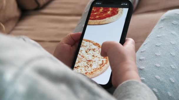自宅でスマホを使ってテイクアウトを注文し クローズアップ 小さな女の子は 携帯電話のアプリケーションを使用してスマートフォン上のピザを選択します 高品質4K映像 — ストック動画