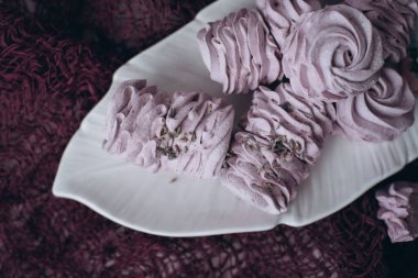 Beyaz bir tabakta yaprak şeklinde lavanta şekerlemeleri. Lavanta çiçekleri serpiştirilmiş marşmelovlar. Tabak mor bir masa örtüsünün üzerinde duruyor. Şekerler. Şekerlemeci