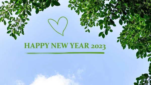 Green text happy new year 2023 with green heart on blue sky and green leaves background concept for greeting and welcoming new year 2023