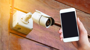 Cep telefonunu tutan bir el, evdeki bir güvenlik kamerasının resmine yerleştirilir. Bu, modern teknolojinin ev güvenliğine entegrasyonunu vurguluyor.