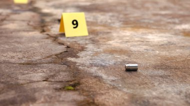 9 numaralı bulanık işaretle yerde yatan boş mermi kovanı. Spor ve güvenlik kavramları hem rekabetçi spor hem de koruyucu tedbirlerde ateşli silahların çifte doğasını vurguluyor.