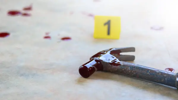 Beton zeminde kan lekeli çelik bir çekiç, 1 numaralı bulanık sarı işaret ve dağınık kan damlaları, soruşturma altındaki suç mahallini sembolize ediyor..