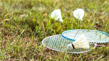 Bir badminton raketi ve bir mekik horozu çimenli bir alanda dinleniyor, açık hava sporlarının neşesini, sağlıklı yaşamı ve spor ve bağ kurmak için eğlence aktivitelerinin önemini sembolize ediyor..