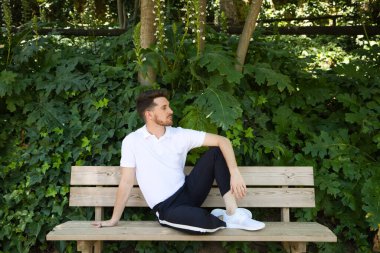 Beyaz tişörtlü yakışıklı genç adam ve siyah pantolon parkta ahşap bankta oturmuş sabırsızlıkla birinin gelmesini bekliyor. Adam farklı vücut ifadeleri kullanıyor. İfade kavramı.