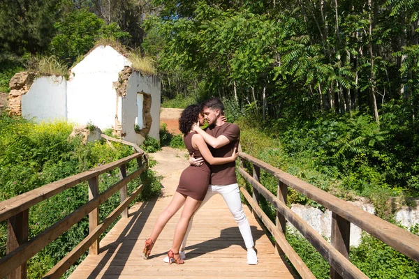 Young and handsome latin dance couple salsa and bachata in the park on a wooden bridge. The couple dances passionately and in love surrounded by greenery and trees. Dance concept.