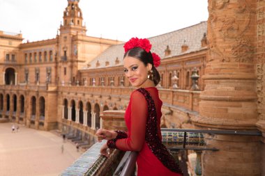 Beautiful teenage woman dancing flamenco on the balcony of a square in Seville. She wears a red dress with ruffles with a lot of art. Flamenco cultural heritage of humanity.