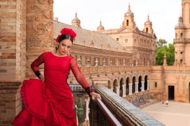 Beautiful teenage woman dancing flamenco on the balcony of a square in Seville. She wears a red dress with ruffles with a lot of art. Flamenco cultural heritage of humanity.