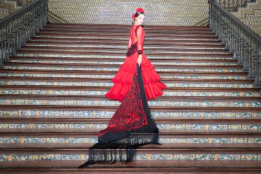 A beautiful teenage woman dancing flamenco goes up the stairs clutching a fringed and embroidered shawl in her hand. She is wearing a red dress with a frill. Flamenco cultural heritage of humanity.