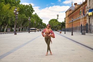 Young, blonde and beautiful woman is on holiday visiting seville. The woman is on a large avenue next to an old palace while walking and posing for photos. Holiday and tourism concept.