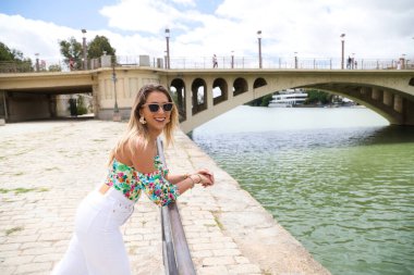 Pretty young blonde woman with sunglasses leaning over the railing of the river. The woman is looking at infinity while smiling or being serious. Various expressions.