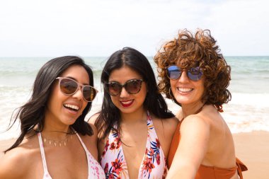 Sahilde arka planda deniz olan üç güzel kadın selfie çekiyor. Kadınlar tatildeler ve fotoğraf için gülümsüyorlar, güneş gözlüğü takıyorlar. Tatil ve seyahat kavramı.