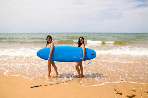 Two young and beautiful women with a surfboard on the shore of the beach. The women are enjoying their trip to the beach paradise. Holidays and travels.