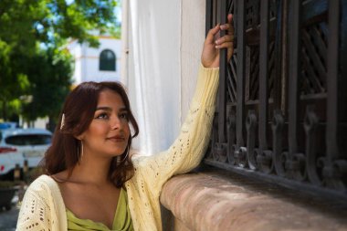 Uzun kıvırcık saçlı ve zarif yeşil bir elbise giyen güzel bir genç kadın. Kadın, Sevilla 'daki eski binanın ızgarasına tutunarak pencerenin yanında duruyor. Tatil kavramı.