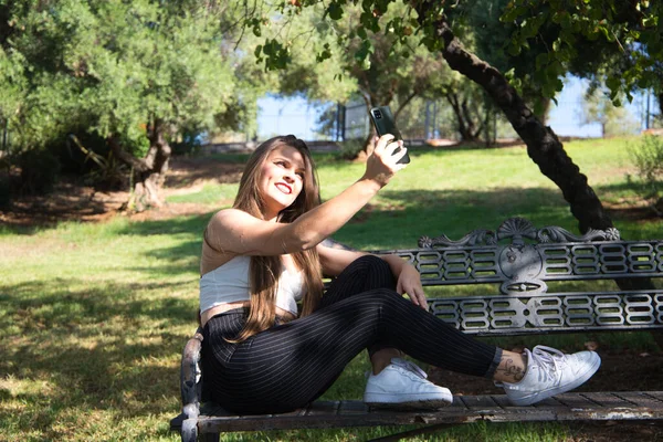 蓝眼睛的女人坐在公园的长椅上 一边拿着手机 一边看社交媒体 一边高兴极了 — 图库照片