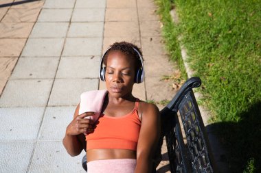 Genç Afro Amerikalı kadın parkta spor yaptıktan sonra bankta oturup omzunda havluyla beyaz kulaklıklarla müzik dinliyor. Spor ve sağlık kavramı.