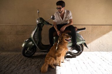 A handsome young man with his brown golden retriever dog. The man is sitting on his retro style motorbike and the dog is sitting on the ground. Concept pets, animals, dogs, pet love, golden retriever.