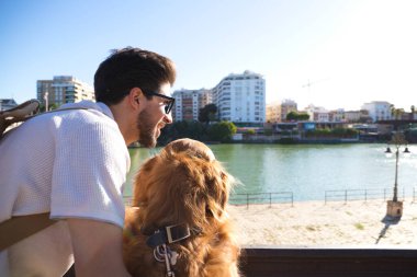 A handsome young man with his brown golden retriever dog leaning on the railing overlooking the river. They are on holiday in seville, spain. Concept pets, animals, dogs, pet love, travel.