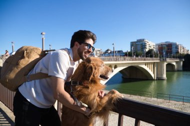 A handsome young man with his brown golden retriever dog leaning on the railing overlooking the river. They are on holiday in seville, spain. Concept pets, animals, dogs, pet love, travel.