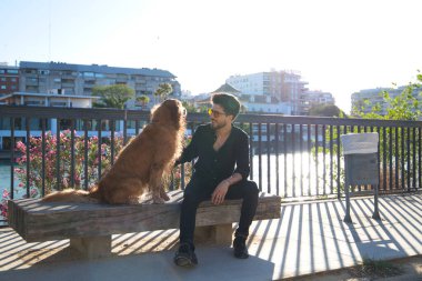 A handsome young man sitting with his golden retriever dog on a bench. In the background you can see the river and against the sunlight in the evening. Concept pets, animals, dogs, pet love.