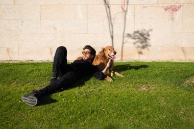 Sakallı ve gözlüklü yakışıklı genç adam kahverengi golden retriever köpeğiyle çimenlerde güneşleniyor. Evcil hayvanlar, hayvanlar, köpekler, evcil hayvanlar, seyahat.