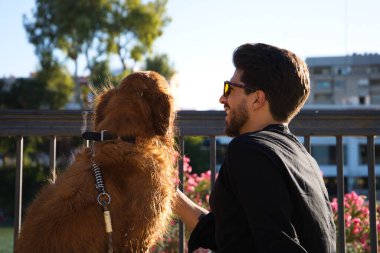 A handsome young man sitting with his golden retriever dog on a bench. In the background you can see the river and against the sunlight in the evening. Concept pets, animals, dogs, pet love.