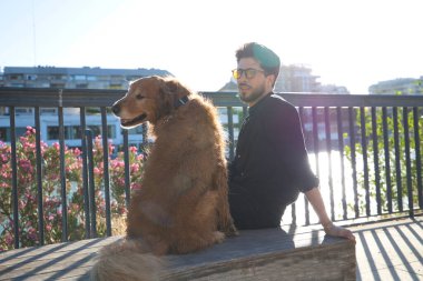 A handsome young man sitting with his golden retriever dog on a bench. In the background you can see the river and against the sunlight in the evening. Concept pets, animals, dogs, pet love.