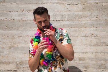 Hawaii çiçekli gömlek, şapka ve renkli çiçek kolyesi giyen yakışıklı genç adam, içinde meşrubat olan bir bardaktan bir kamışdan su içiyor. Tatil ve seyahat kavramı.