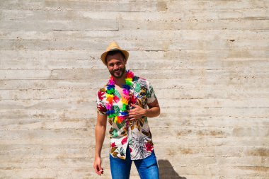 Havai çiçekli gömlek, şapka ve renkli çiçekli kolye giyen yakışıklı bir genç. Adam çok mutlu. Arka plan gri duvar. Seyahat ve tatil konsepti.