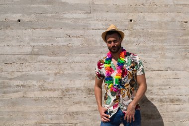 Havai çiçekli gömlek, şapka ve renkli çiçekli kolye giyen yakışıklı bir genç. Adam çok mutlu. Arka plan gri duvar. Seyahat ve tatil konsepti.