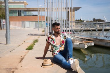 Yakışıklı genç adam Hawaii çiçekleri ve şapkası olan bir gömlek giyiyor ve iskele güneşlenirken nehir kıyısında oturuyor. Tatil ve boş zaman ve seyahat konsepti.