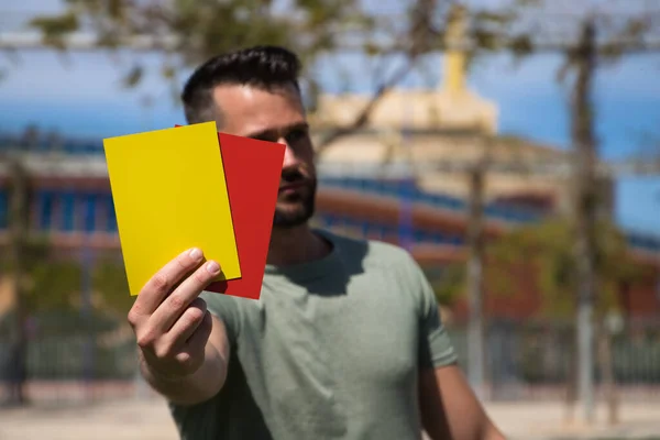 stock image Handsome young man holding a football in his hand while showing red and yellow card to camera as if he were a football referee. Concept of sports and team play.