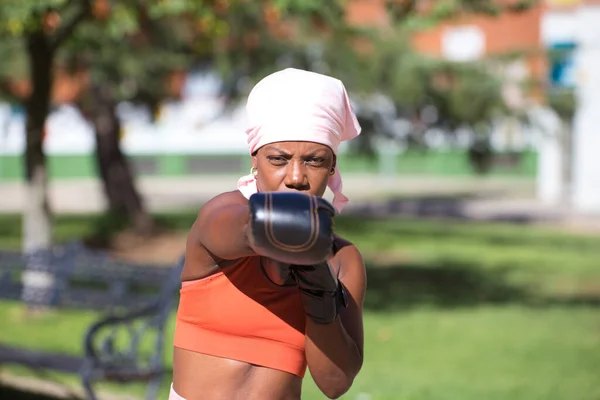 Afro American woman playing sports in the park. The woman has boxing gloves and a pink cancer scarf on her head. Concept of breast cancer and health.