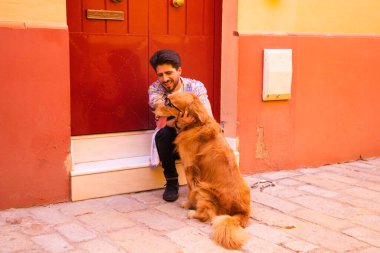 Genç, yakışıklı İspanyol bir adam, kahverengi köpeğiyle, bir basamakta dinleniyorlar, arka planda kırmızı bir kapı. Evcil hayvanlar, hayvanlar, köpekler, evcil hayvanlar, Golden Retre