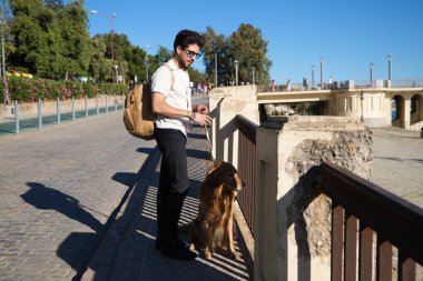 A handsome young man with his brown golden retriever dog leaning on the railing overlooking the river. They are on holiday in seville, spain. Concept pets, animals, dogs, pet love, travel.