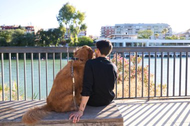 A handsome young man sitting with his golden retriever dog on a bench. In the background you can see the river and against the sunlight in the evening. Concept pets, animals, dogs, pet love.