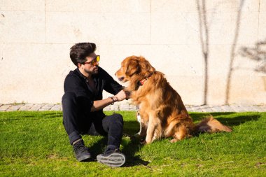 Sakallı ve gözlüklü yakışıklı genç adam çimenlerde oturmuş köpeğiyle güneşleniyor. Evcil hayvanlar, hayvanlar, köpekler, evcil hayvanlar, seyahat.