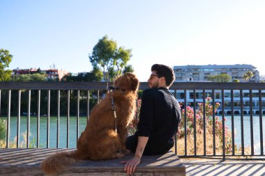A handsome young man sitting with his golden retriever dog on a bench. In the background you can see the river and against the sunlight in the evening. Concept pets, animals, dogs, pet love.