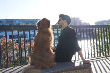 A handsome young man sitting with his golden retriever dog on a bench. In the background you can see the river and against the sunlight in the evening. Concept pets, animals, dogs, pet love.