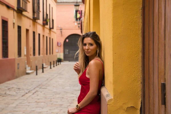 Young, blonde, beautiful woman in a red dress is visiting seville. The woman poses for the camera very elegant and like a model in the typical streets of the city. Holidays and travels