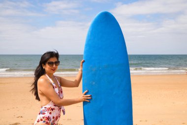 Mavi sörf tahtası olan genç ve güzel bir Latin kadın. Kadın kumsalda. Tatil ve yaz konsepti.