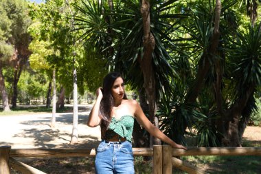 Parktaki korkuluklara yaslanmış siyah saçlı güzel bir kadın. Kadın fotoğraflara poz verirken farklı vücut pozları veriyor. Sonbahar ve doğa kavramı.