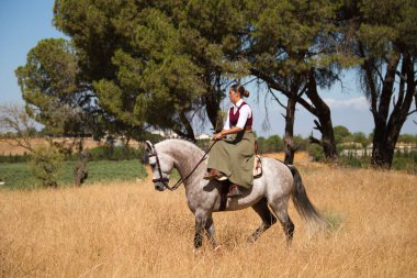 Young and beautiful Spanish woman on a Thoroughbred horse riding in the countryside in Spain. The woman is wearing a horse riding uniform. Thoroughbred and equine concept.