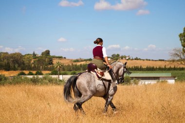 Young and beautiful Spanish woman on a Thoroughbred horse riding in the countryside in Spain. The woman is wearing a horse riding uniform. Thoroughbred and equine concept.