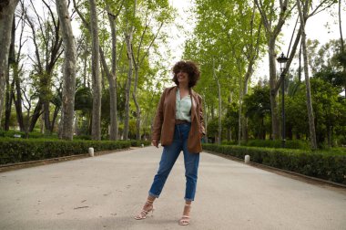 Orta yaşlı, güzel, kıvırcık saçlı İspanyol bir kadın bir Avrupa şehrinde büyük ağaçları olan bir parkta duruyor. Kadın modern ve modaya uygun kıyafetler giyiyor. Seyahat ve tatil konsepti.