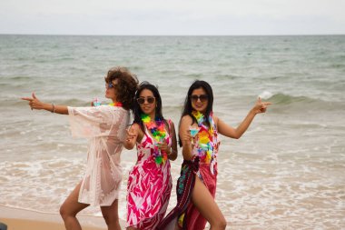 three young and beautiful latin women with a glass of blue cocktail stroll along the beach happily talking to each other and having fun. Holiday and travel concept.